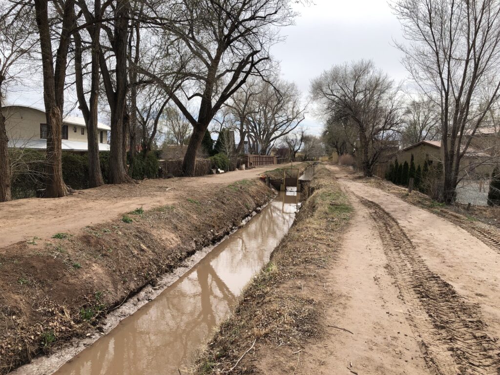 Irrigation ditch flanked by bare dirt roads and trees with no leaves. A small amount of brown water in the ditch.