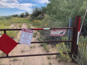 Gate and "no trespassing" signs blocked a sandy dirt road, with lush green vegetation to the right and an irrigation ditch to the left