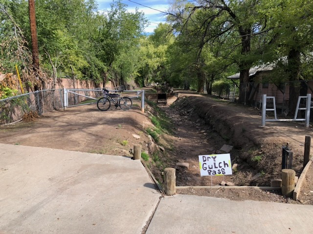 A dry irrigation ditch flanked by green trees with a hand-made sign reading "Dry Gulch Pass." Bike leaning against ditch bank gate.