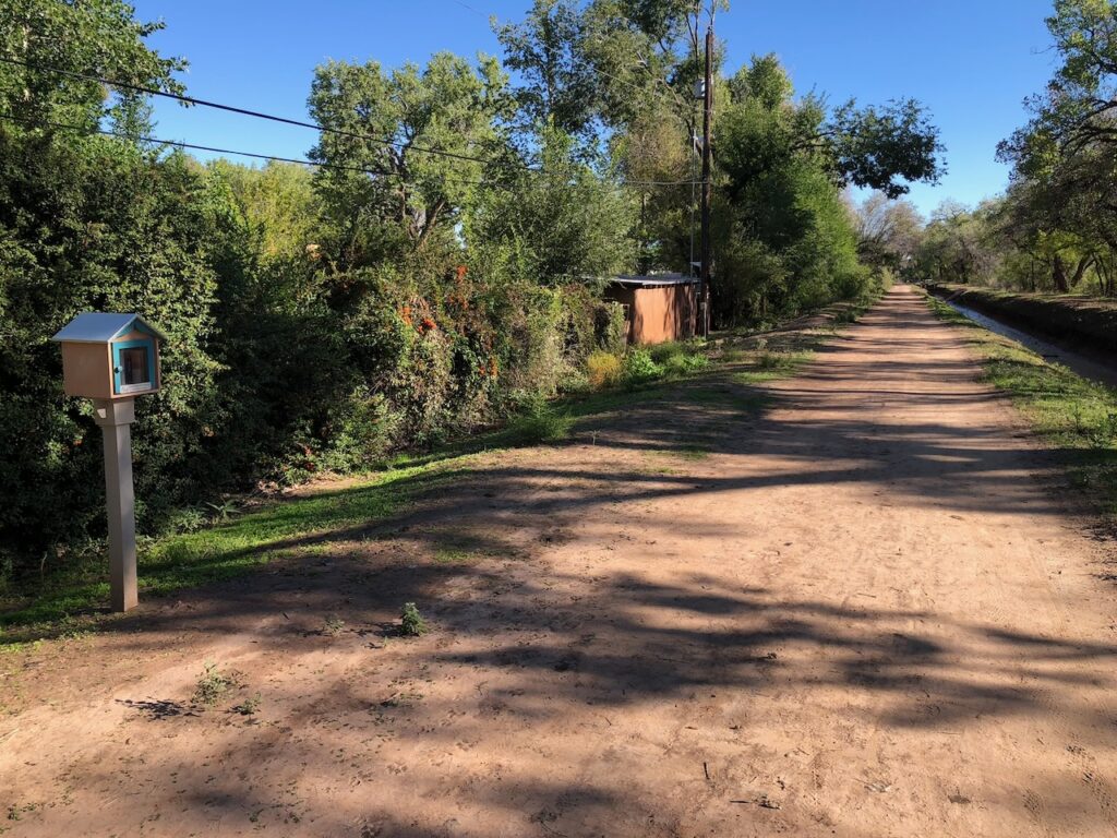 Shaded dirt path along an irrigation ditch with a Little Free Library book sharing box to one side.