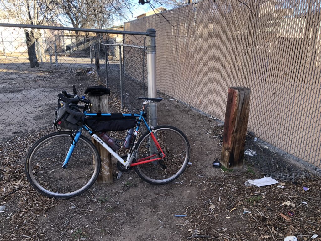 A blue and red touring bicycle with bags attached to the frame and handlebars is parked against a wooden post in an urban setting. The area is strewn with dry leaves and some litter on the ground. There's a chain-link fence in the background with a concrete post beside the bike. The setting appears to be a neglected urban corner with no people in sight.