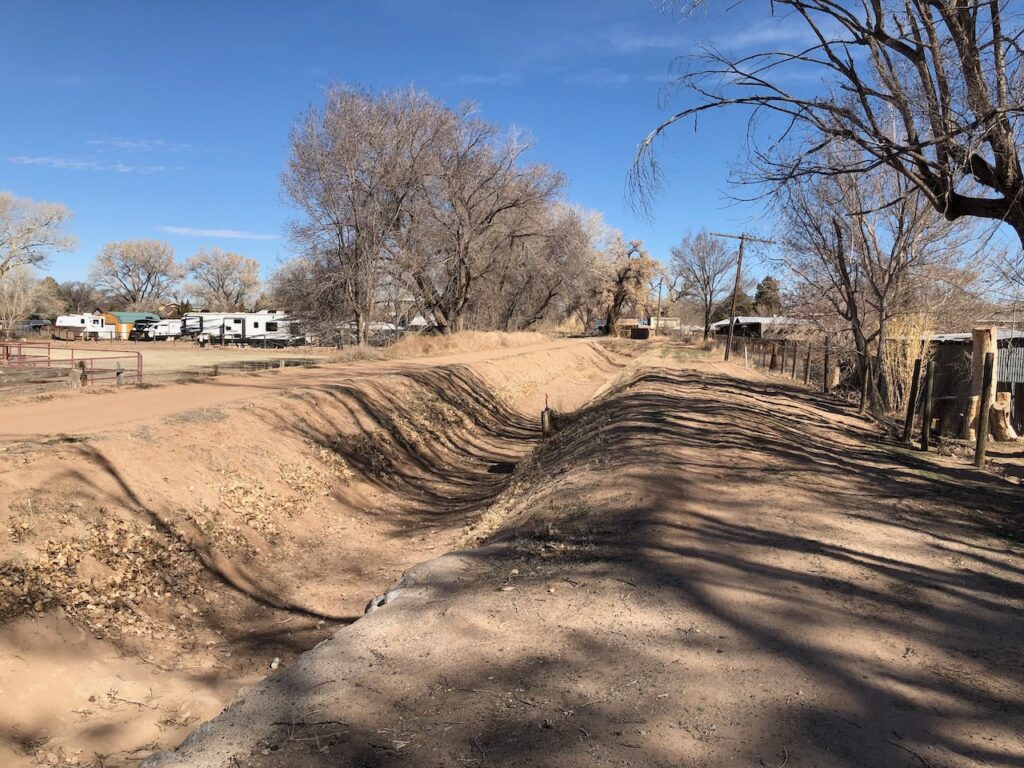 A dry irrigation ditch, brown, lined with trees and homes and a horse corral