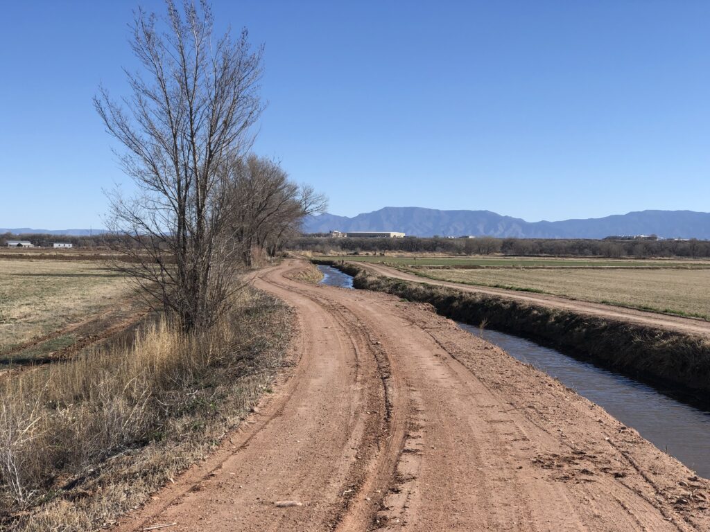 A winding irrigation ditch with a dirt road, trees, and mountains in the background, surrounded by farm fields not yet green.