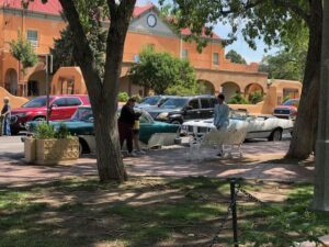 beautifully restored classic cars parked on a tree-lined street, with a historic adobe church and lush greenery in the background.