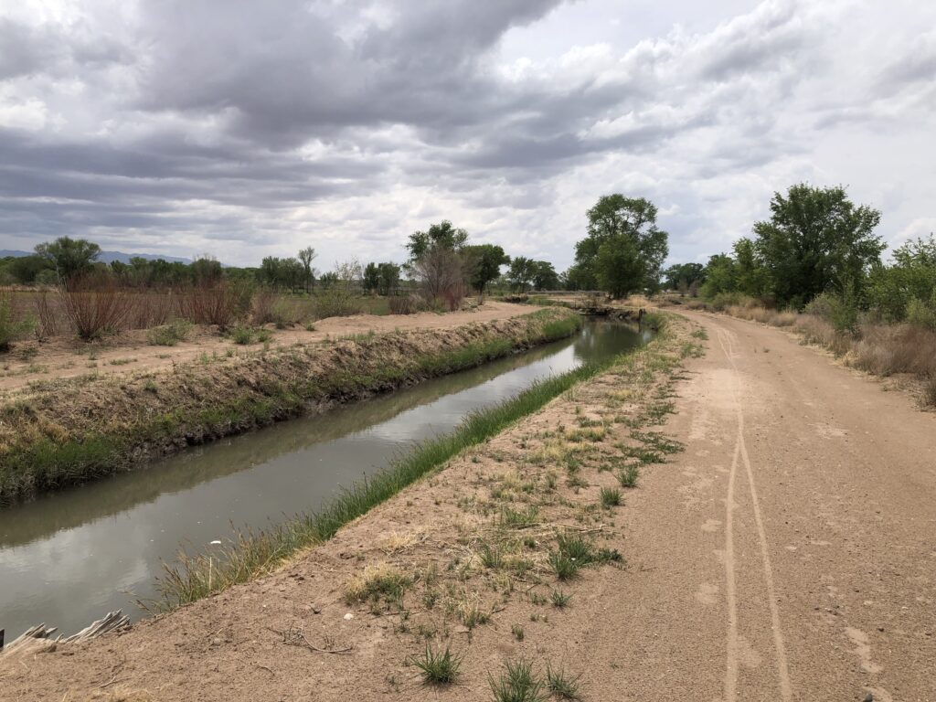 An irrigation ditch flanked by a dirt road with trees and a cloudy sky.