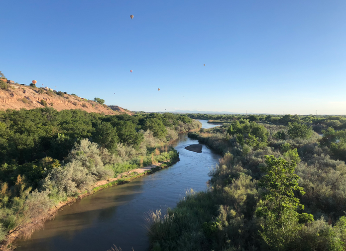 River flanked by woods with desert bluff to the left and tiny hot air balloons dotting the sky