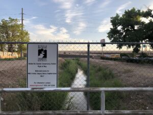 Canal with water passing beneath a freeway with motel to the right and "Middle Rio Grande Conservancy District" sign in the foreground.